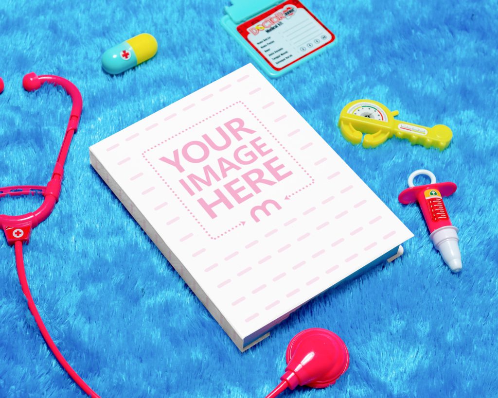 A book on a blue furry background surrounded by a toy stethoscope, syringe, note paper and pill.