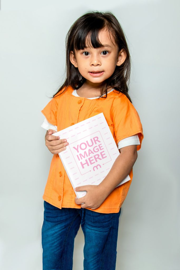 A little girl is standing and holding a book. Holds the book with both hands. He is standing with a white background behind him.