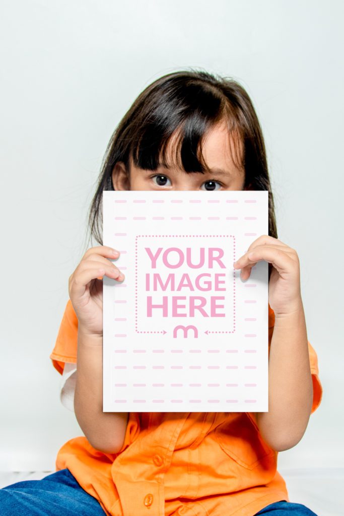 A cute little girl with black hair is holding a book in front of her face. He is sitting with a white background behind him.