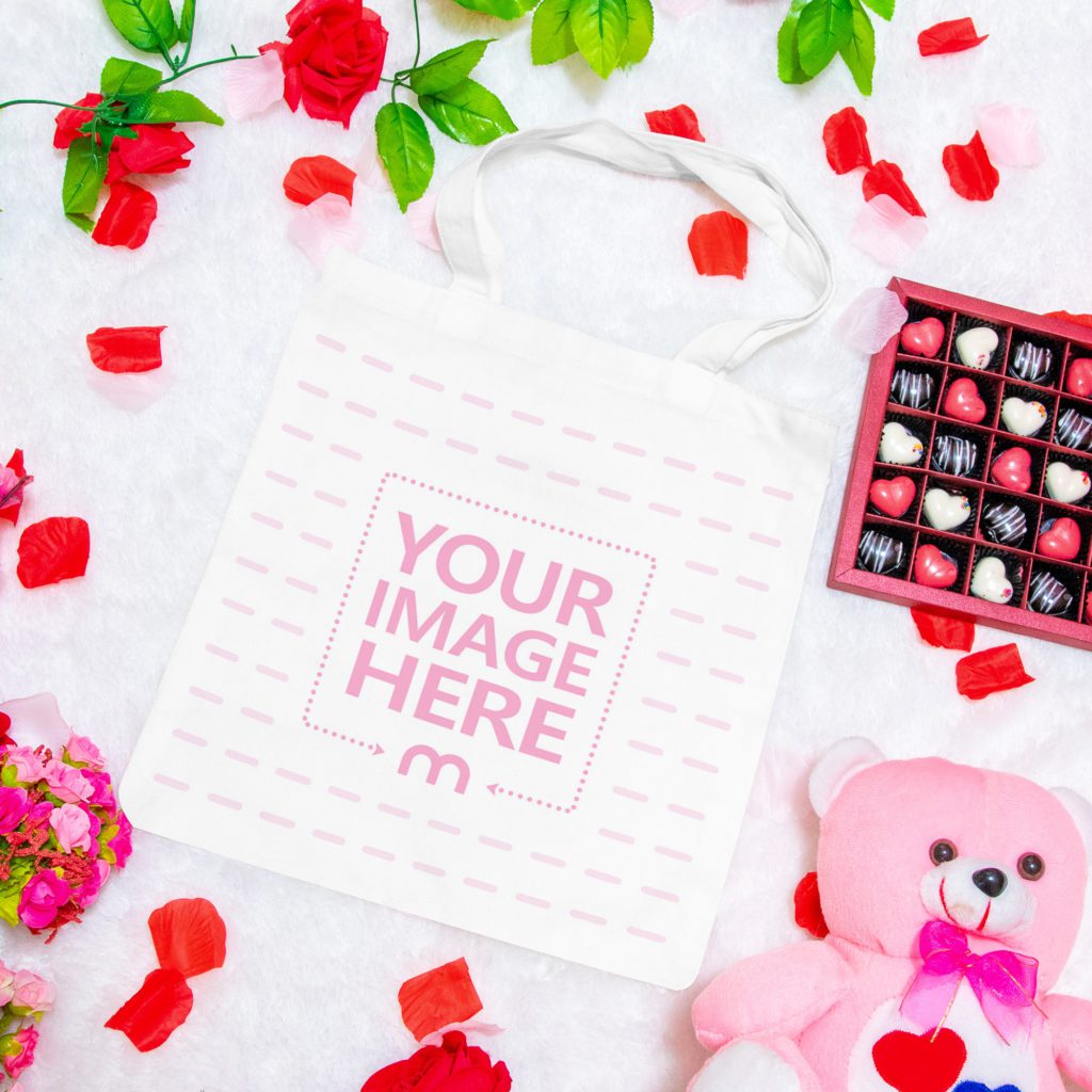 A tote bag on a white background. Around him are heart-shaped chocolate candies, a teddy bear and rose flowers.