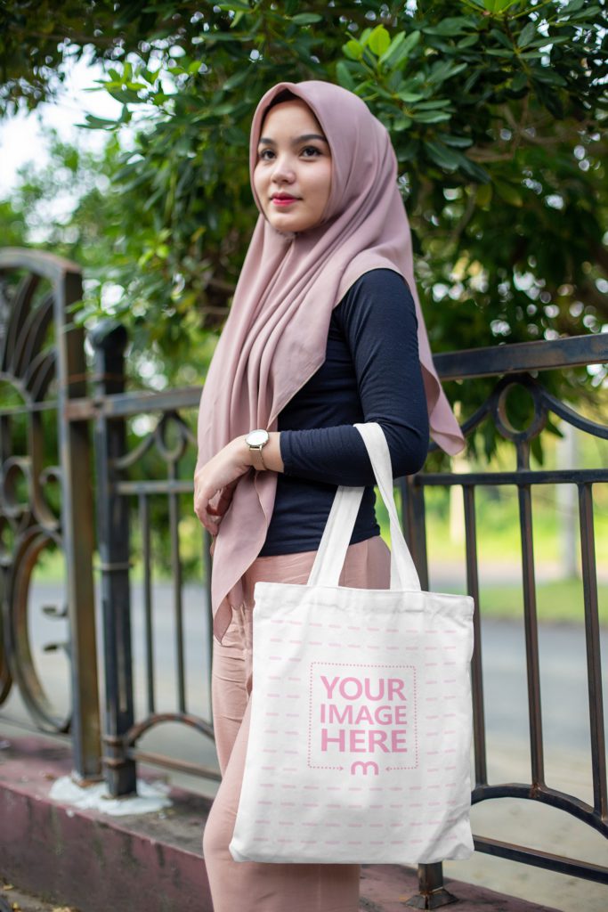 Muslim woman posing outside with a tote bag. In his background is a garden and a tree. The woman is smiling and the bag is on her hand.